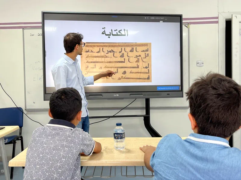 Teacher at a smartboard displaying an early Kufic Qur'an manuscript, two students seated in front