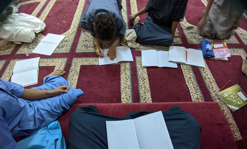 Children sitting on a patterned carpet with open notebooks, practising Arabic writing